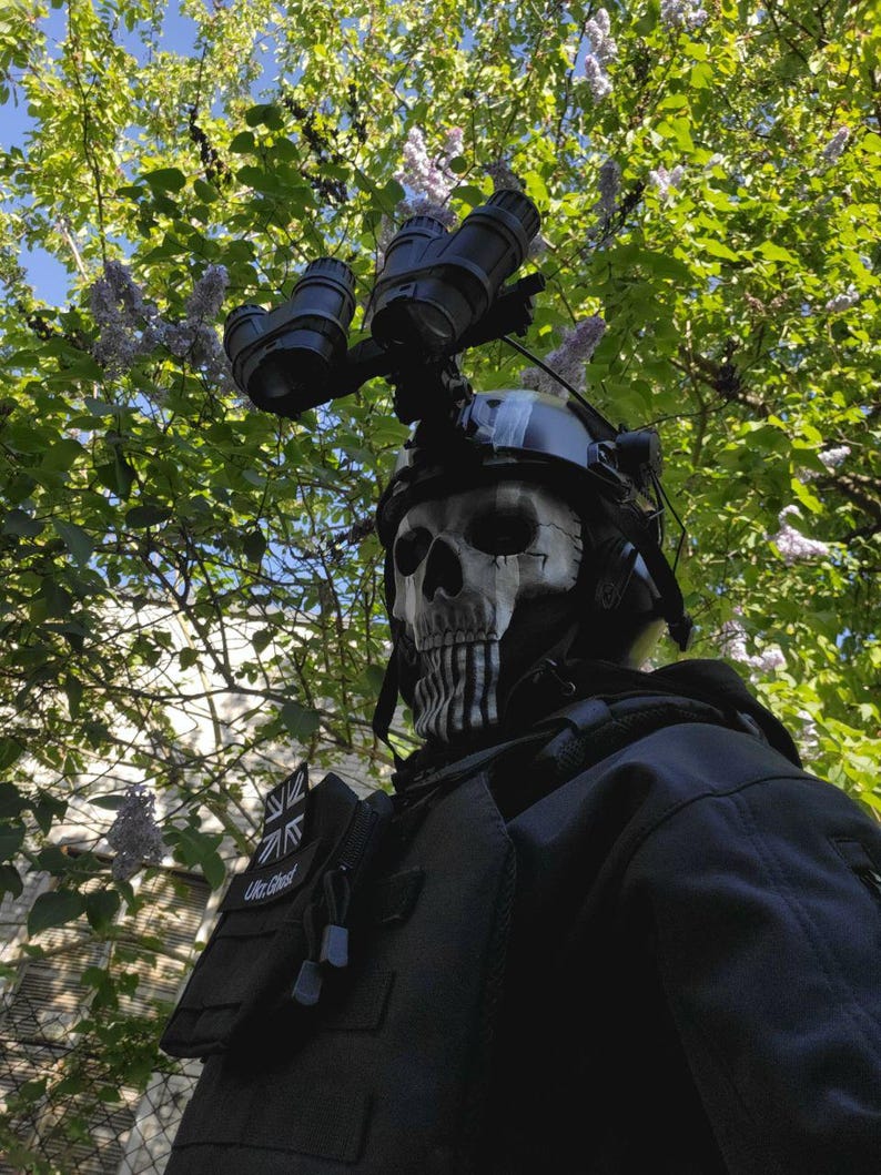 以下が含まれることがあります： 黒いタクティカルヘルメットに暗視ゴーグルとスカルマスク。マスクは黒いアクセントが付いた白いスカルデザインです。その人は「UK Ghost」のパッチが付いた黒いタクティカルベストを着ています。