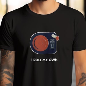 Könnte beinhalten: Schwarzes T-Shirt mit einer Grafik einer Vintage-Filmrolle und dem Text "I ROLL MY OWN". Das Design umfasst eine rote Rolle, ein blau-weißes Gehäuse und einen roten Knopf. Das T-Shirt hat einen klassischen Rundhalsausschnitt.