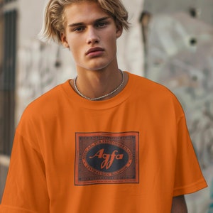 Puede incluir: Camiseta naranja brillante con un diseño rectangular que muestra la palabra "Agfa" en una fuente estilizada. El diseño está enmarcado por un borde rojo con detalles en blanco y negro. La camiseta tiene mangas cortas y un corte holgado.
