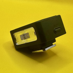 Puede incluir: Un flash de cámara rectangular negro con una lente transparente y estriada. La lente tiene un patrón circular en el interior. Una base de metal plateado está unida en la parte inferior. El flash está sobre un fondo amarillo.