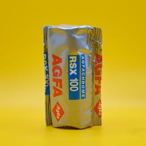 May include: A silver Agfa RSX 100 film roll against a yellow background. The film packaging has the Agfa logo and the text "RSX 100" in a blue rectangle. The Agfa logo is also in red.