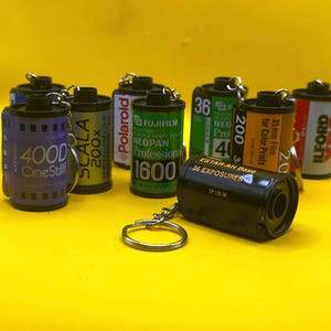 Puede incluir: Una colección de mini carretes de película con llaveros. Los carretes presentan varias marcas y tipos de película, incluyendo CineStill, Scala, Fujifilm e Ilford. Los colores varían entre negro, verde, amarillo y morado, sobre un fondo amarillo brillante.