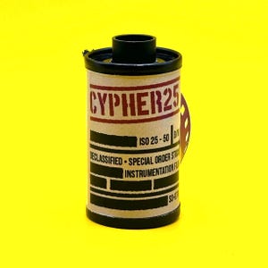 May include: A cylindrical film canister with the text "CYPHER25" in red letters. The canister has a black top and bottom, with a tan body. Additional text includes "ISO 25-50 B/W", "DECLASSIFIED - SPECIAL ORDER STOCK", and "INSTRUMENTATION FILM".