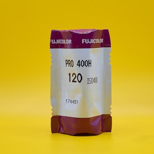 Puede incluir: Un paquete de película Fujicolor Pro 400H 120, con un diseño morado y blanco. El paquete de película tiene el texto "PRO 400H", "120" e "ISO400" impreso. El fondo es amarillo liso.