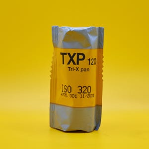 Puede incluir: Un rollo de película TXP 120 Tri-X pan, con un envoltorio amarillo y plateado. La película tiene una ISO de 320, y el embalaje incluye la fecha 11/2001. El fondo es amarillo.