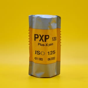 Puede incluir: Un rollo de película PXP 120 Plus-X pan, envuelto en un paquete plateado y amarillo. La etiqueta amarilla muestra el texto "PXP 120", "Plus-X pan" e "ISO 125". La película está sobre un fondo amarillo brillante.