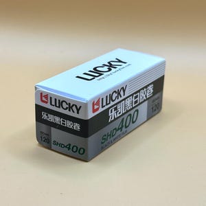 May include: A white, black, and grey box of Lucky SHD400 black and white film. The box features the Lucky logo, Chinese characters, and the text "SHD400" and "120". The film is designed for 120 format cameras.