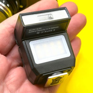 May include: A black camera flash with a silver hot shoe mount. The flash has a rectangular shape with a white light panel and a small information panel that reads "Model: SL03 Battery Capacity: 3.7V 300mAh". The flash is held in a hand.
