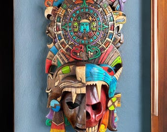 Puede incluir: Un colgante de pared de madera tallada que presenta un diseño detallado de calendario azteca sobre una máscara dividida. La máscara es mitad marrón y mitad roja, con intrincadas tallas y acentos coloridos. La pieza está montada en una pared azul.