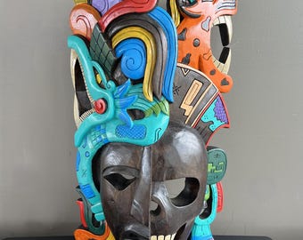 Puede incluir: Una máscara de madera tallada con un diseño de media calavera y figuras de animales estilizadas y coloridas. La máscara presenta una calavera marrón oscuro en la mitad inferior, con una serpiente turquesa y un jaguar naranja arriba. La máscara está pintada con colores brillantes.