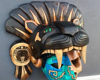 Puede incluir: Una máscara de madera tallada con un tocado de jaguar con ojos verdes y grandes colmillos. La máscara tiene una cara azul y turquesa con diseños geométricos y una figura de madera tallada en el lateral. La máscara está montada en una pared gris.