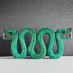 Op de afbeelding: Een groene, slangachtige sculptuur met een gebarsten, getextureerd oppervlak. De sculptuur heeft twee gestileerde slangenkoppen met open monden, die tanden en rode accenten onthullen. Het is een decoratief item.