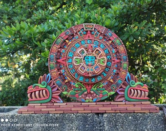 May include: A colorful wooden replica of the Aztec calendar, known as the Sun Stone, with intricate carvings and vibrant colors. The calendar is mounted on a brick-like base with two green and red serpent heads on either side.