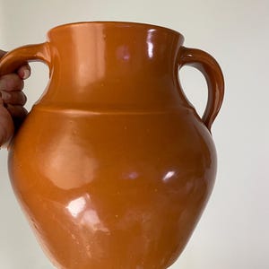 May include: A glazed, orange-brown ceramic pitcher with two curved handles. The pitcher has a rounded body and a wide mouth. The surface is smooth and reflective, with subtle variations in color. This rustic vessel is suitable for decorative or functional use.