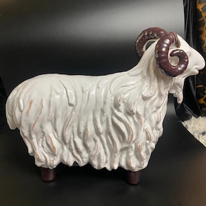 May include: A white ceramic ram sculpture with brown horns and hooves. The ram has a textured, shaggy coat and a detailed face. The sculpture is a decorative item, likely for home decor.
