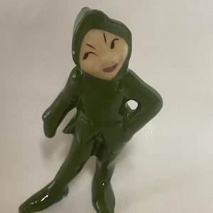 May include: A small, green figurine of a character in a hooded outfit, with a smiling face and arms akimbo. The figurine is standing with one leg extended, and the details are painted in a glossy finish.