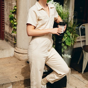 May include: A person wearing a beige short-sleeved jumpsuit with a collared neckline and a zipper. The person is wearing sunglasses and white sneakers. They are holding a black-lidded coffee cup. The outfit is casual and stylish.