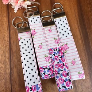 Puede incluir: Cuatro llaveros de tela con herrajes metálicos. Los llaveros presentan varios patrones, incluyendo blanco con lunares negros, rayas rosas y diseños florales con flores rosas y azules. Un arreglo floral rosa está en el fondo.