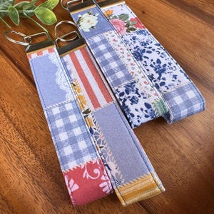 Puede incluir: Cuatro llaveros de tela con anillas metálicas. Cada llavero presenta un diseño de patchwork con estampados florales, de cuadros y a rayas en tonos azules, rojos y amarillos. Los llaveros están sobre una superficie de madera.