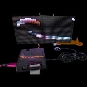 May include: A black electronic device with a screen displaying a pixelated game. The device has a black and purple box with the text "ROTOFRENZ" and a button. A pink USB cable is connected to the box. The screen shows a game with white, blue, and red pixels.