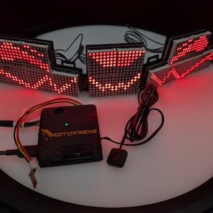 May include: A set of red LED matrix displays arranged in a semi-circle, displaying animated patterns. A black control module with the text "ROTOFRENZ" is connected to the displays. The setup is on a white, circular surface.