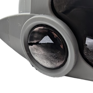 May include: A close-up of a grey, futuristic device featuring a circular, black lens. The lens has a reflective surface with a triangular shape in the centre. The device appears to be made of a matte, light grey material.