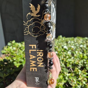 May include: A beaded bookmark with pink, orange, black, and grey beads, and a gold-coloured charm. The bookmark is in a book titled "Iron Flame". The book has a black cover with gold lettering and a dragon graphic.