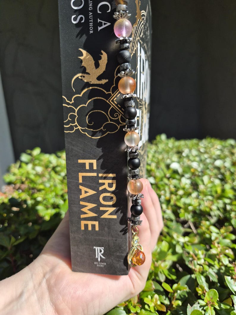 May include: A beaded bookmark with pink, orange, black, and grey beads, and a gold-coloured charm. The bookmark is in a book titled "Iron Flame". The book has a black cover with gold lettering and a dragon graphic.