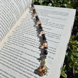 May include: A beaded bookmark with a gold-coloured charm and a gemstone pendant. The beads are black, peach, and clear, strung together with silver-coloured accents. The bookmark is placed on an open book.