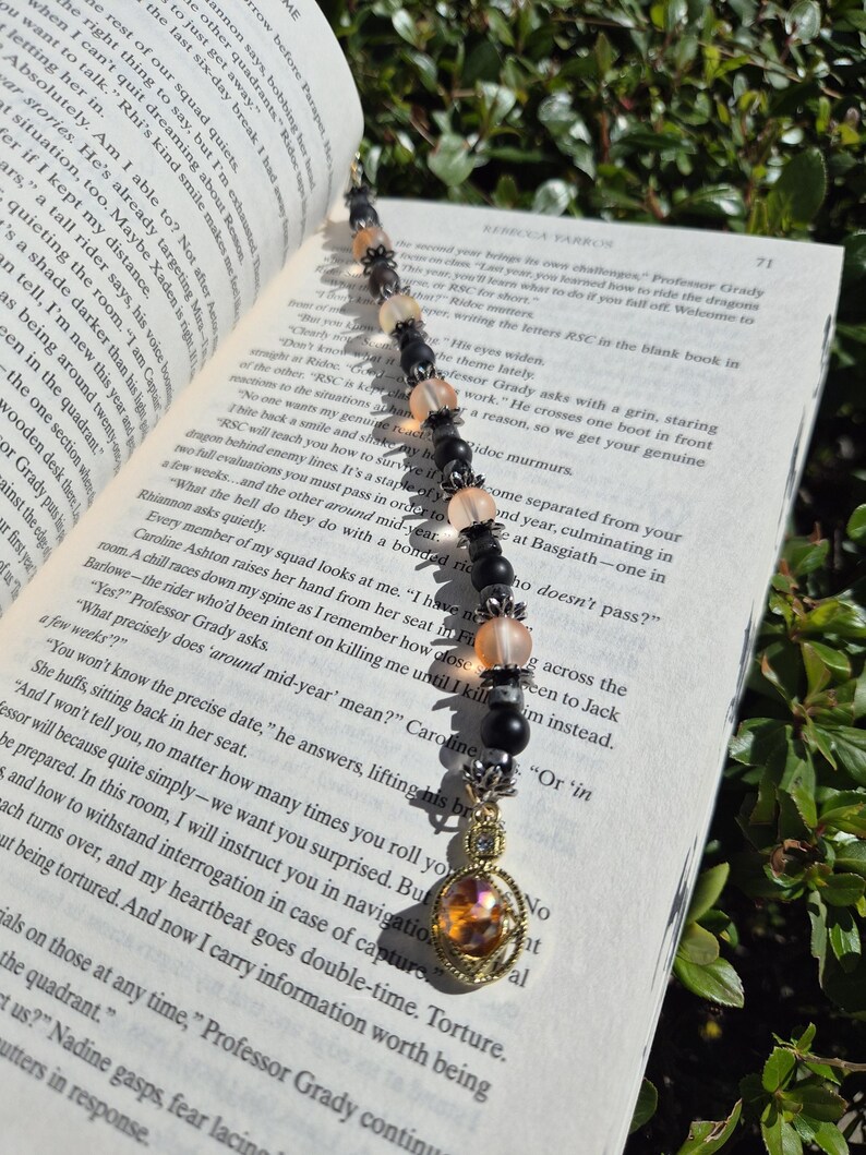 May include: A beaded bookmark with a gold-coloured charm and a gemstone pendant. The beads are black, peach, and clear, strung together with silver-coloured accents. The bookmark is placed on an open book.