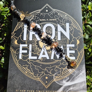 May include: A beaded bookmark lies on a book titled "Iron Flame" by Rebecca Yarros. The bookmark has black and peach-coloured beads, a gold-coloured charm, and a small metal dragon. The book cover is black with gold lettering and design.