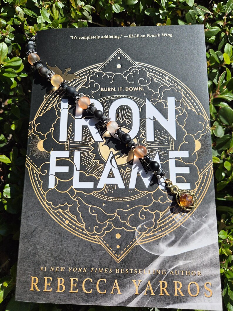 May include: A beaded bookmark lies on a book titled "Iron Flame" by Rebecca Yarros. The bookmark has black and peach-coloured beads, a gold-coloured charm, and a small metal dragon. The book cover is black with gold lettering and design.