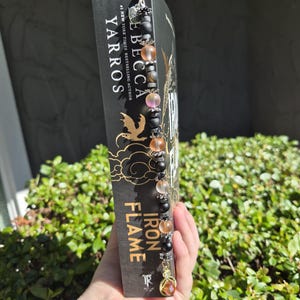 May include: A beaded bookmark with a gold charm is attached to a book titled "Iron Flame" by Rebecca Yarros. The bookmark features black, clear, and orange beads. The book has a black cover with gold lettering and a gold dragon design.