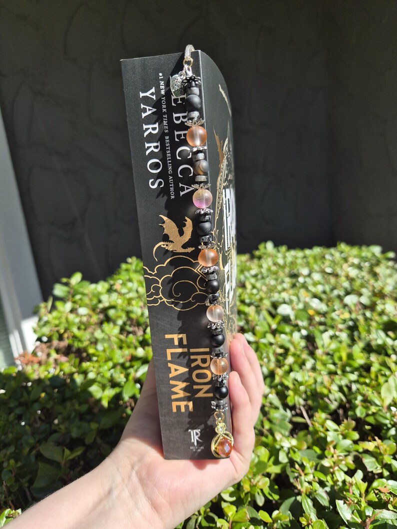 May include: A beaded bookmark with a gold charm is attached to a book titled "Iron Flame" by Rebecca Yarros. The bookmark features black, clear, and orange beads. The book has a black cover with gold lettering and a gold dragon design.