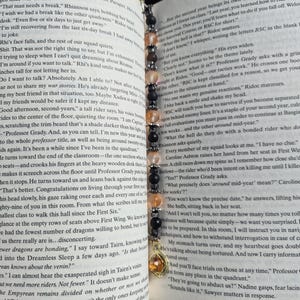 May include: A beaded bookmark with a gold charm, placed in a book. The bookmark has black, peach, and clear beads. The book is open, with text visible.