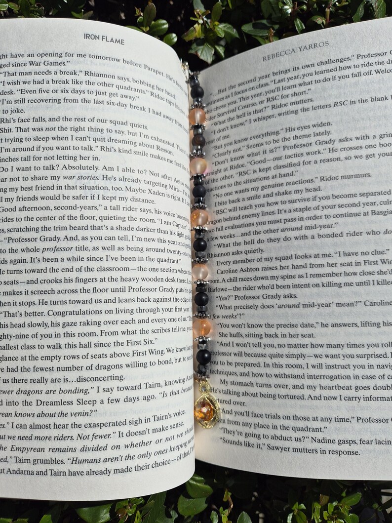 May include: A beaded bookmark with a gold charm, placed in a book. The bookmark has black, peach, and clear beads. The book is open, with text visible.