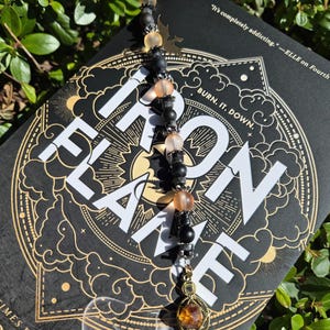 May include: A beaded bookmark with black, peach, and gold beads, and a gold-coloured charm. The bookmark is resting on a book with the title "Iron Flame" and the text "BURN. IT. DOWN."
