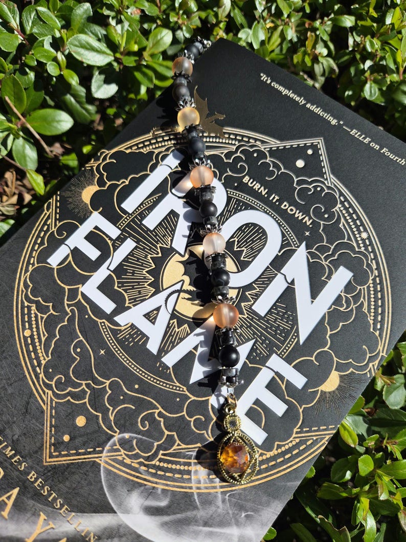 May include: A beaded bookmark with black, peach, and gold beads, and a gold-coloured charm. The bookmark is resting on a book with the title "Iron Flame" and the text "BURN. IT. DOWN."