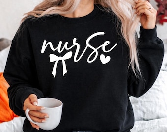 Puede incluir: Sudadera negra con la palabra "nurse" en escritura cursiva blanca, un lazo y un corazón. El texto está centrado en la parte delantera de la parte superior de manga larga. La persona sostiene una taza blanca.