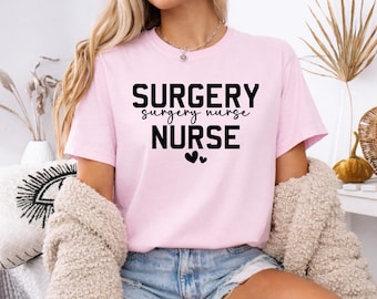 Puede incluir: Camiseta rosa claro con la frase "SURGERY surgery nurse NURSE" en letras negras. Dos pequeños corazones negros están debajo de la palabra "NURSE". La camiseta es de manga corta.