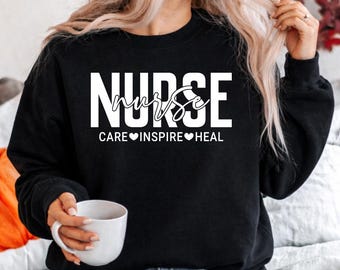 Puede incluir: Sudadera negra con la palabra "NURSE" en letras blancas grandes. La palabra "nurse" está escrita en cursiva encima. Debajo, las palabras "CARE INSPIRE HEAL" con símbolos de corazón.