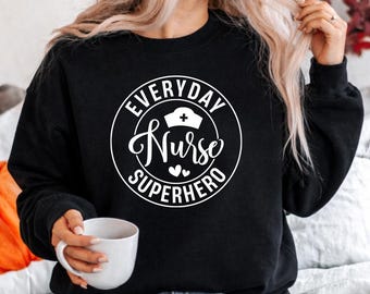 Puede incluir: Sudadera negra con un diseño circular blanco que dice "Everyday Nurse Superhero". El diseño incluye un gorro de enfermera y corazones. Una taza blanca se sostiene en la mano.