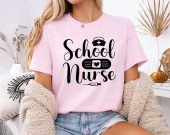 Puede incluir: Camiseta rosa claro con la frase "School Nurse" en negro. El diseño incluye un gorro de enfermera, una tirita con un corazón y una jeringuilla. La camiseta es de tejido suave.