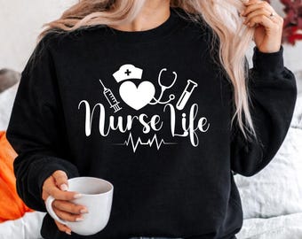 Puede incluir: Sudadera negra con la frase blanca "Nurse Life". El diseño incluye un gorro de enfermera, un corazón, un estetoscopio, una jeringa y un vial. Una taza blanca se sostiene en la mano.