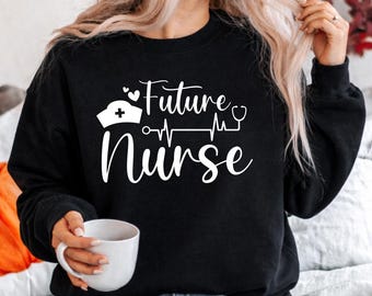 Puede incluir: Sudadera negra con el texto blanco "Future Nurse". El diseño incluye un gorro de enfermera, una línea de latidos y un estetoscopio. Una taza blanca se sostiene en la mano.