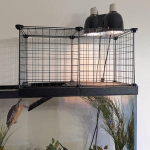 May include: A black wire mesh basking platform for an aquarium, featuring a rectangular cage structure with two black dome heat lamps mounted on top. The platform sits atop a glass tank containing water, aquatic plants, and a swimming turtle.