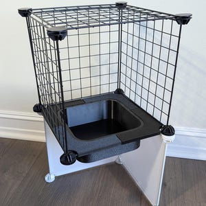 May include: A modular pet enclosure featuring a black wire grid frame with four top corner connectors. The structure includes a central black basin with a metallic interior, supported by white plastic panels and white corner connectors at the base.