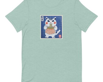 Puede incluir: Una camiseta azul claro con una ilustración de estilo vintage de un gato blanco comiendo fideos. El gato lleva un chaleco estampado en rojo y blanco y la ilustración está enmarcada en un borde blanco. El texto japonés "猫飯" está impreso sobre el gato.