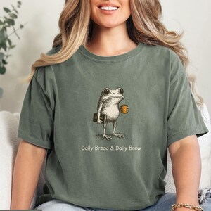 May include: Moss green t-shirt featuring a whimsical frog illustration holding a book and a coffee mug. The text "Daily Bread & Daily Brew" is printed below the frog. The shirt is a relaxed fit.