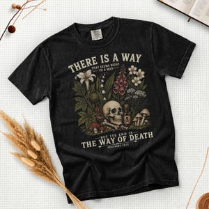May include: A black short-sleeve t-shirt featuring a graphic print of a human skull, various wildflowers, mushrooms, and a small bottle with a skull and crossbones label. The text reads: THERE IS A WAY THAT SEEMS RIGHT TO A MAN BUT ITS END IS THE WAY OF DEATH PROVERBS 14:12.